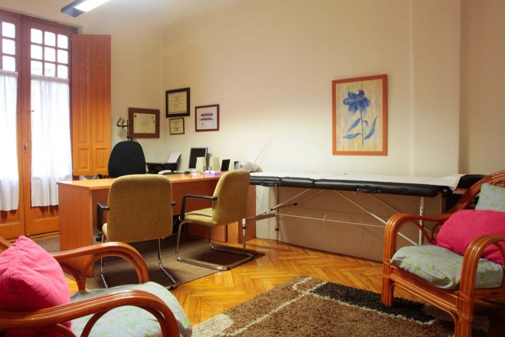 Oficina con escritorio, sillas y camilla de terapia. Suelo de madera, ventana con luz. Arte en las paredes, color neutro.