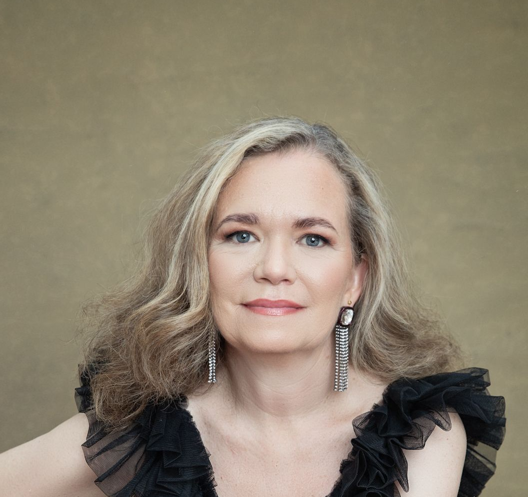 Mujer de cabello rubio grisáceo con un top negro con volantes y pendientes de plata colgantes. Posa sobre un fondo dorado.