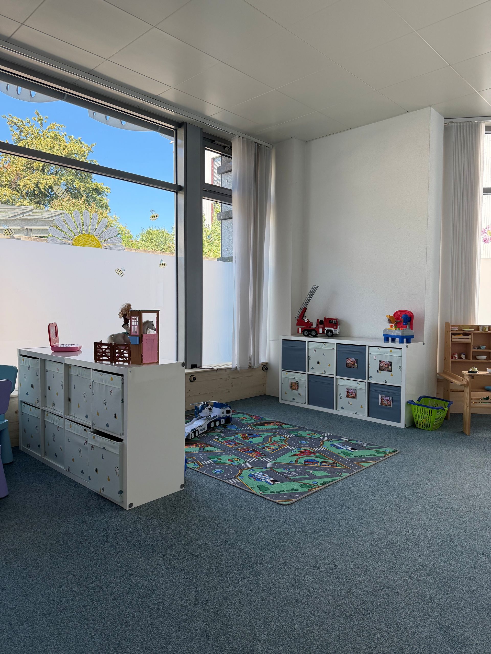 Ein Erwachsener und ein Kleinkind spielen in einem hellen Spielzimmer mit einem bunten Spielzeug; im Hintergrund ist ein weiteres Kind zu sehen.