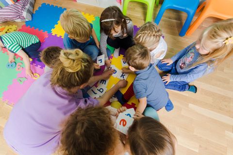 Auf dem bunten Klassenzimmerboden versammeln sich Kinder um eine Lehrerin und spielen mit Spielzeug und Bauklötzen.
