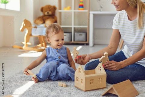 Kleinkind spielt in einem hellen Wohnzimmer mit einem Spielzeughaus auf dem Boden, während ein Erwachsener ihm hilft.