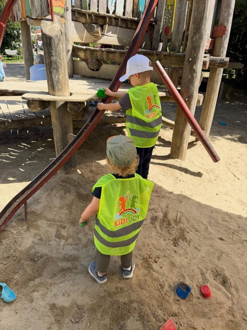 Zwei Kinder in Warnwesten klettern auf ein hölzernes Spielgerät auf einem sandigen Spielplatz.