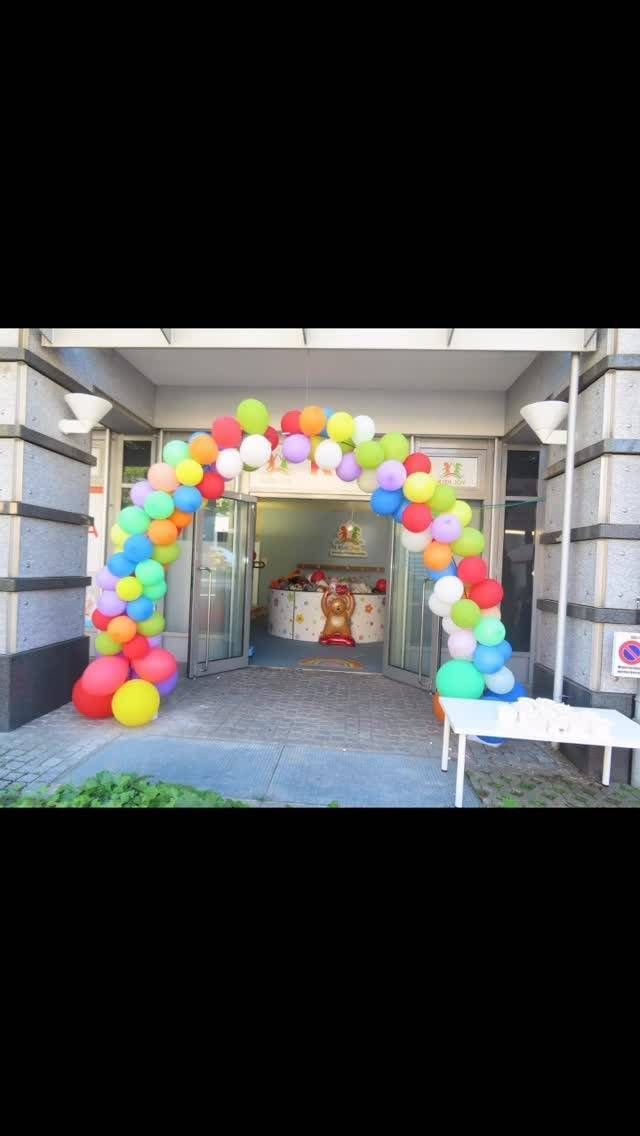 Ein bunter Ballonbogen über dem Eingang eines Gebäudes, rechts davon ein Tisch, von dem aus man die dekorierte Inneneinrichtung sehen kann.