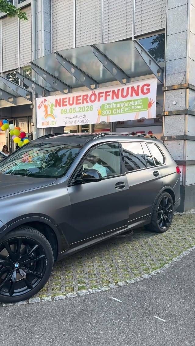 Ein mattschwarzer SUV parkt vor einem Ladenlokal mit einem Banner zur Eröffnung „NEUERÖFFNUNG“.