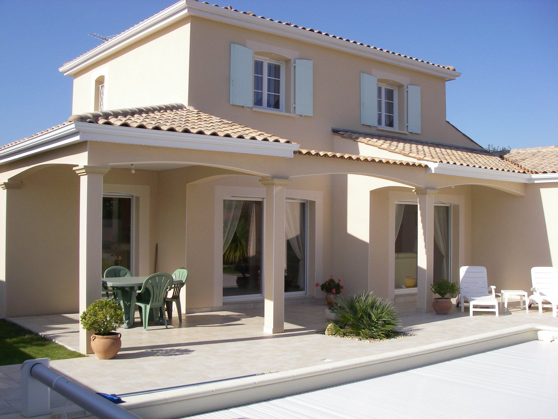 Maison à étage avec façade claire, volets bleu pâle et patio couvert avec piscine.
