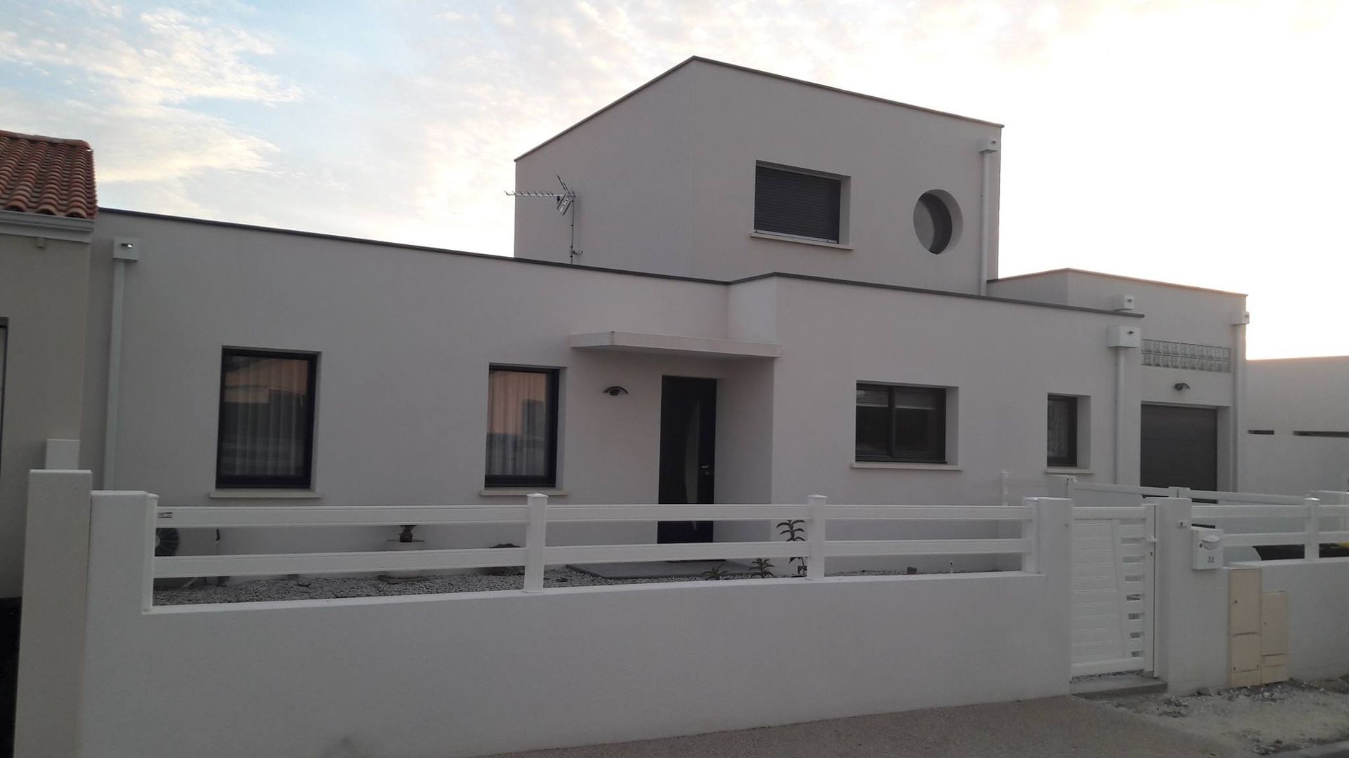 Maison blanche à deux étages avec des fenêtres noires, une fenêtre ronde et une clôture blanche.