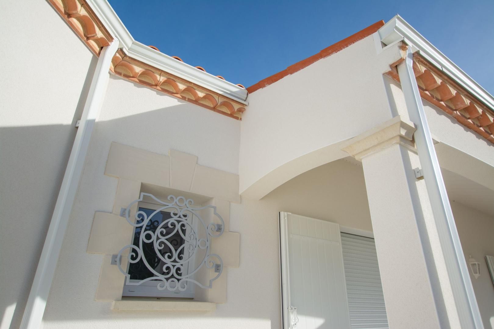 Angle d'un bâtiment blanc avec une fenêtre ornée et une toiture en tuiles de terre cuite.