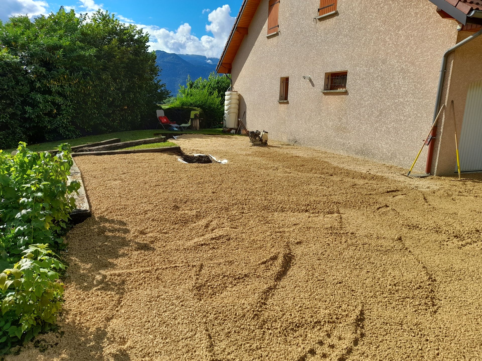 Une maison avec du gravier beige dans la cour, une montagne et des buissons visibles en arrière-plan par une journée ensoleillée.