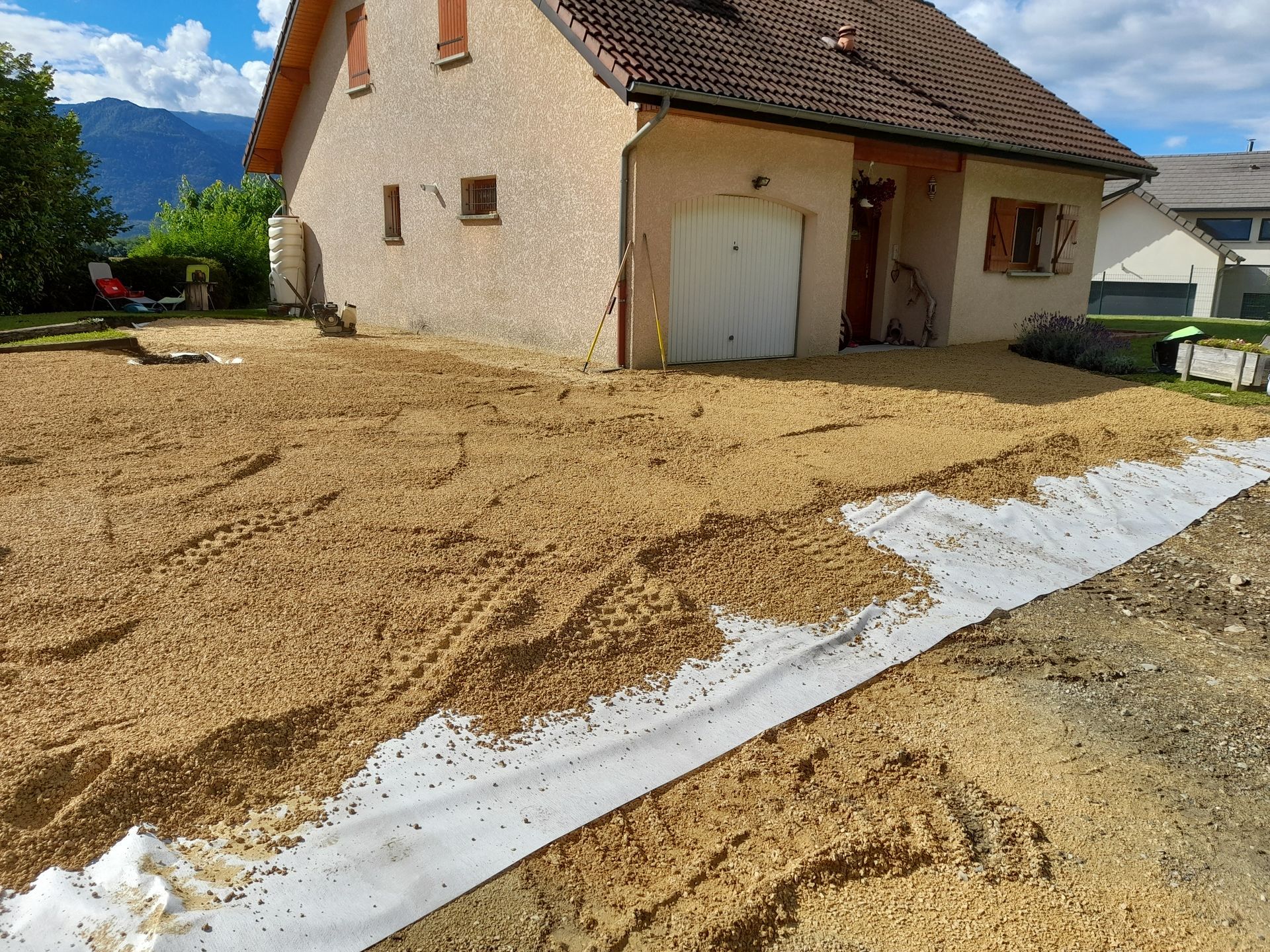 Extérieur d'une maison avec une cour recouverte de gravier, toile de paillage blanche, montagnes en arrière-plan.