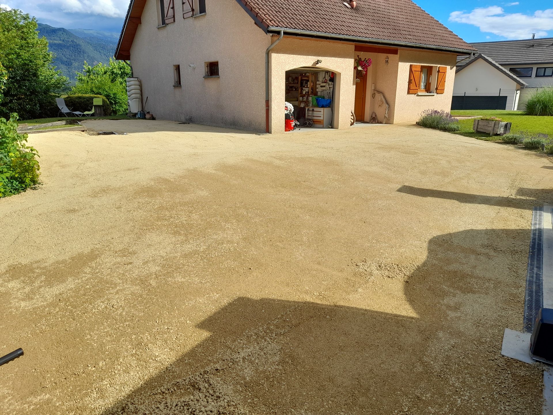 Allée de gravier devant une maison beige avec une porte rouge et des fenêtres à volets, sous un ciel nuageux.
