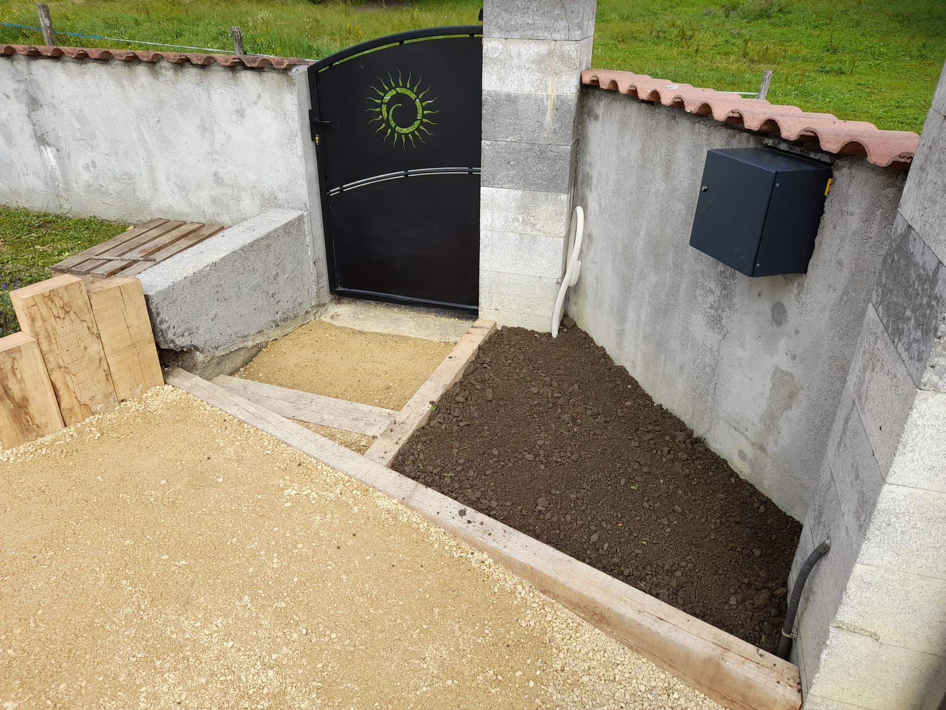 Un portail noir orné d'un soleil, des marches en gravier et une boîte aux lettres adossée à un mur.