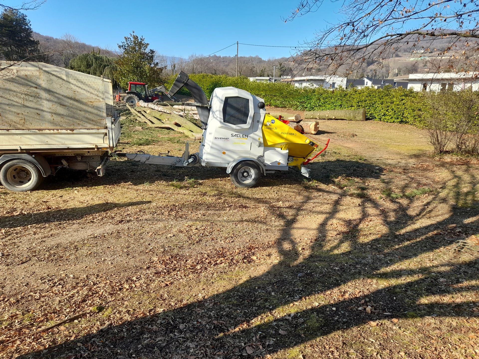 Un petit camion blanc avec un attelage jaune est stationné sur un terrain en terre battue, avec une remorque. On aperçoit des montagnes en arrière-plan.