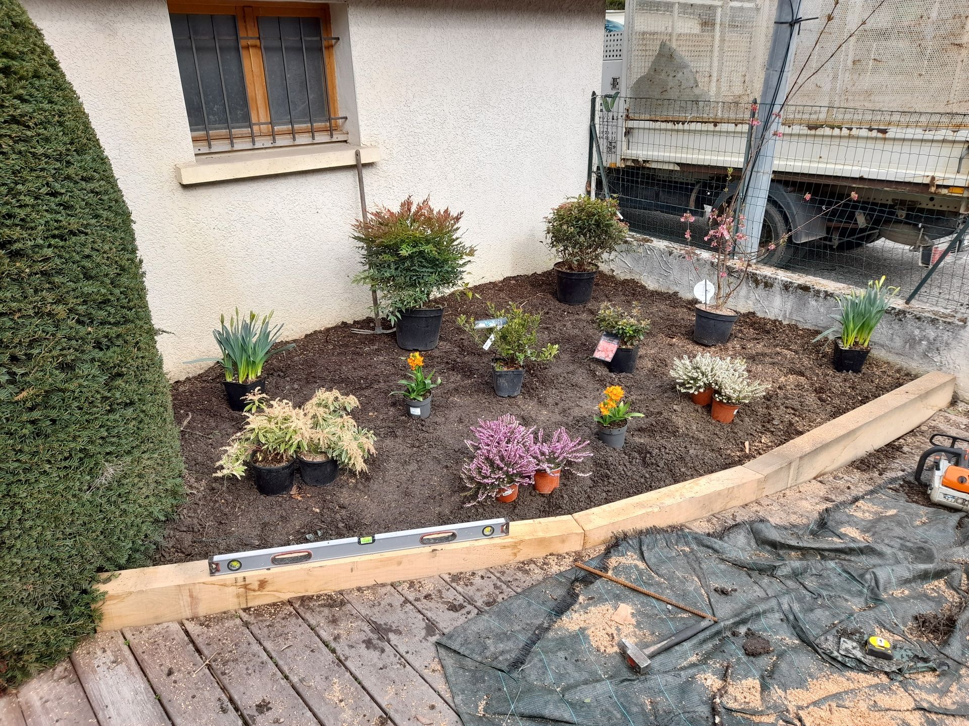 Un parterre de fleurs avec des plantes en pots et du paillis, bordé de bois. Un bâtiment blanc se dresse en arrière-plan.