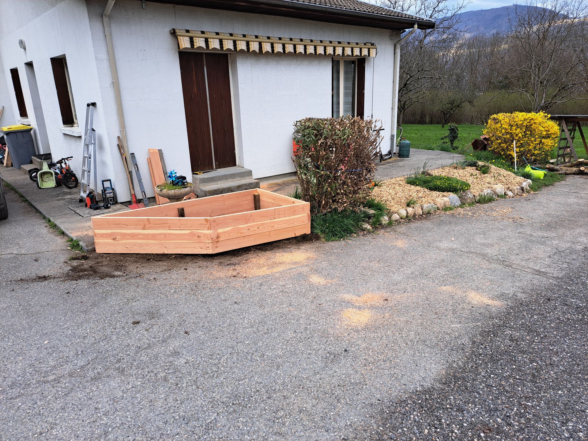 Jardinière en bois sur une allée de gravier devant une maison, aménagement paysager à droite.