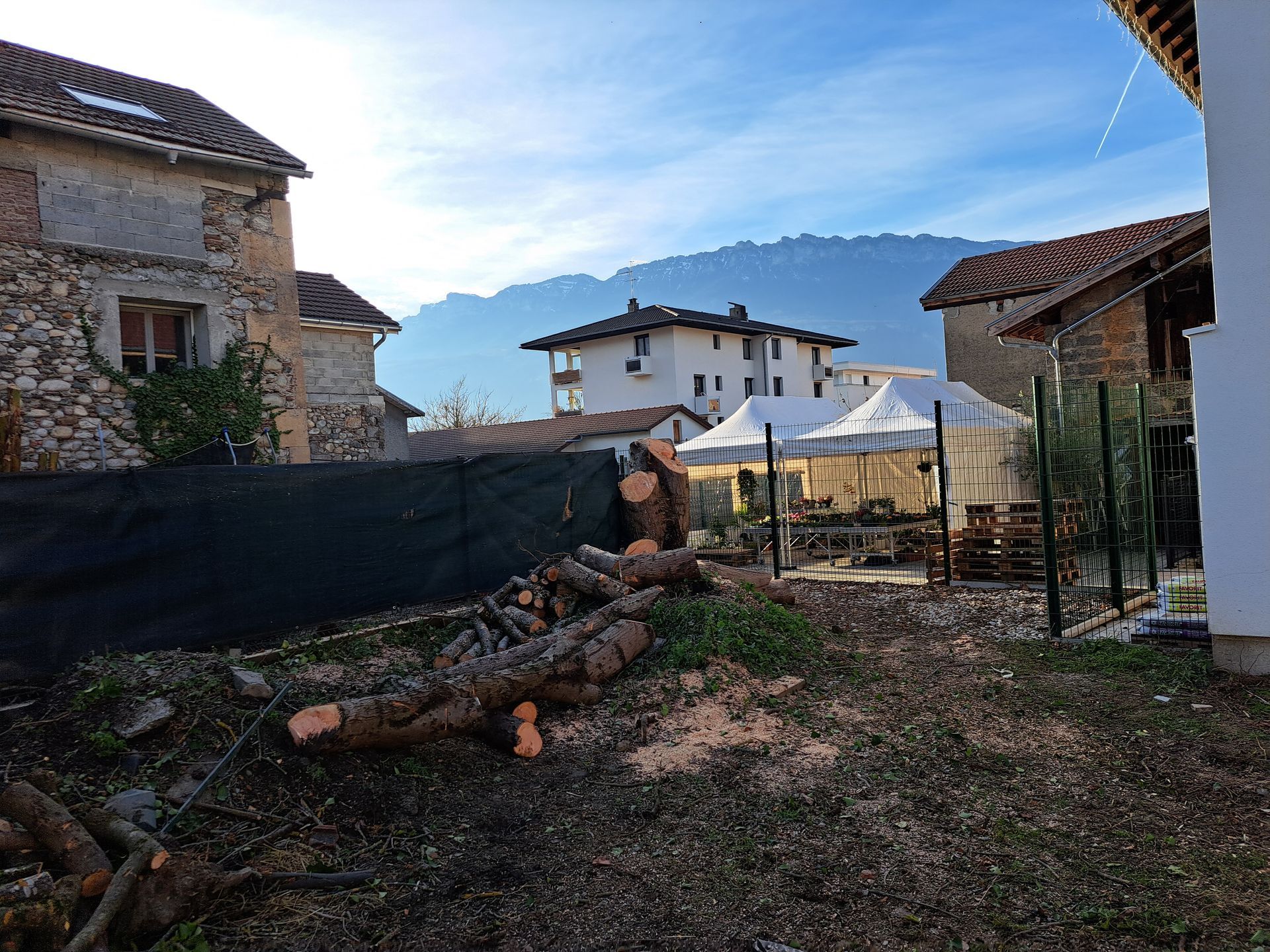 Des bûches et des débris jonchent une cour, avec des bâtiments et des montagnes en arrière-plan, sous un ciel bleu.