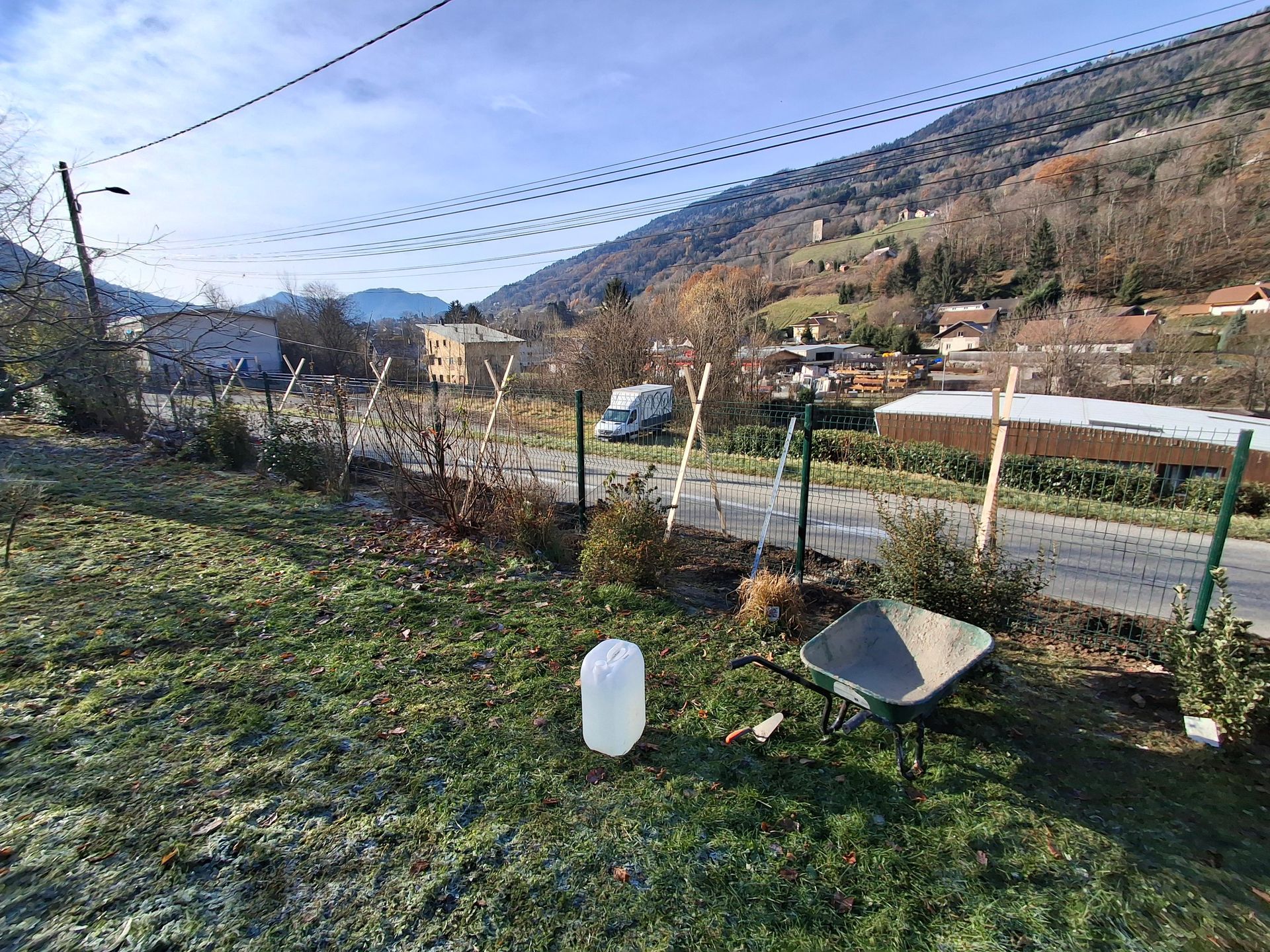 Pelouse avec une brouette, un bidon d'eau et une route, avec des montagnes et des bâtiments en arrière-plan.