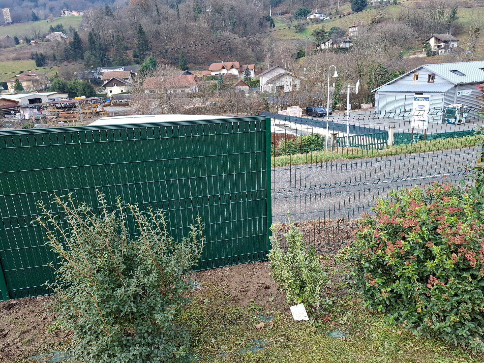 Au premier plan, une clôture verte agrémentée de plantes. À l'arrière-plan, des bâtiments et des arbres à flanc de colline.