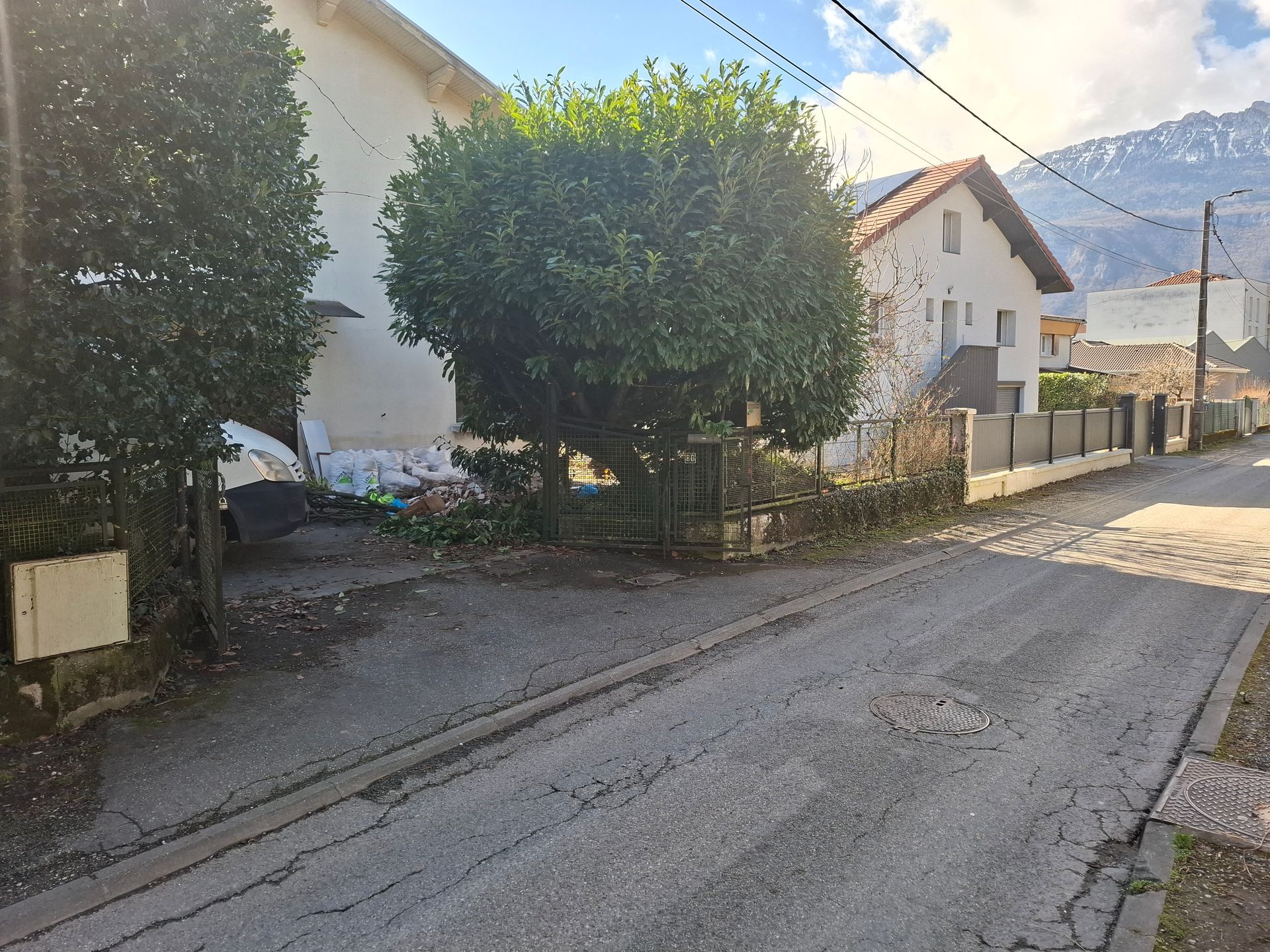 Rue avec des maisons, des arbres et une montagne en arrière-plan sous un ciel lumineux.