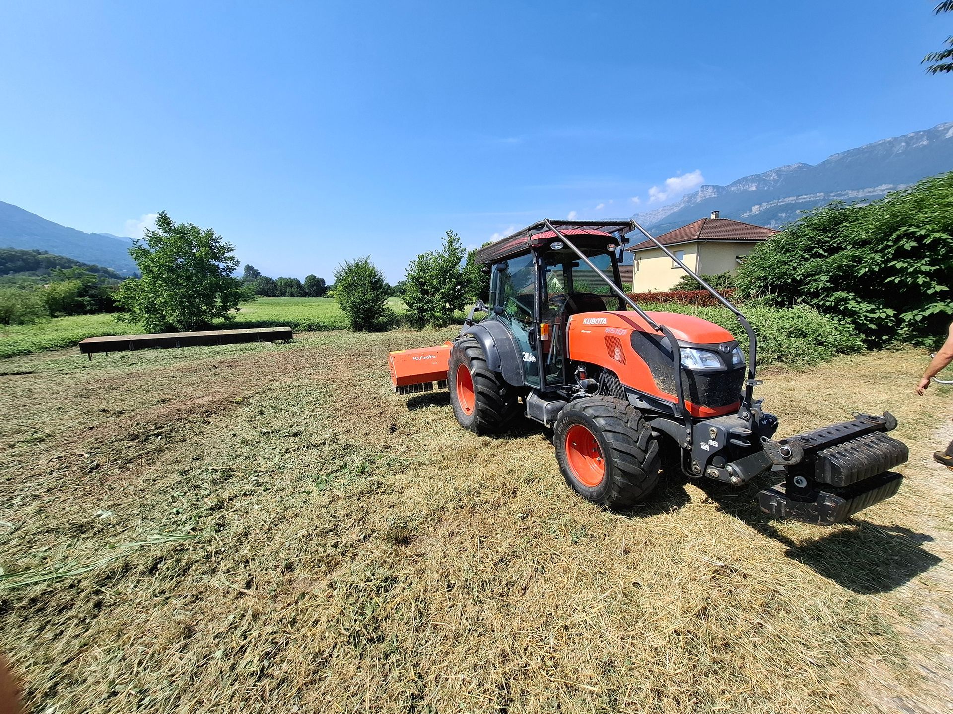 Tracteur orange et gris dans un champ sous un ciel bleu, montagnes en arrière-plan.