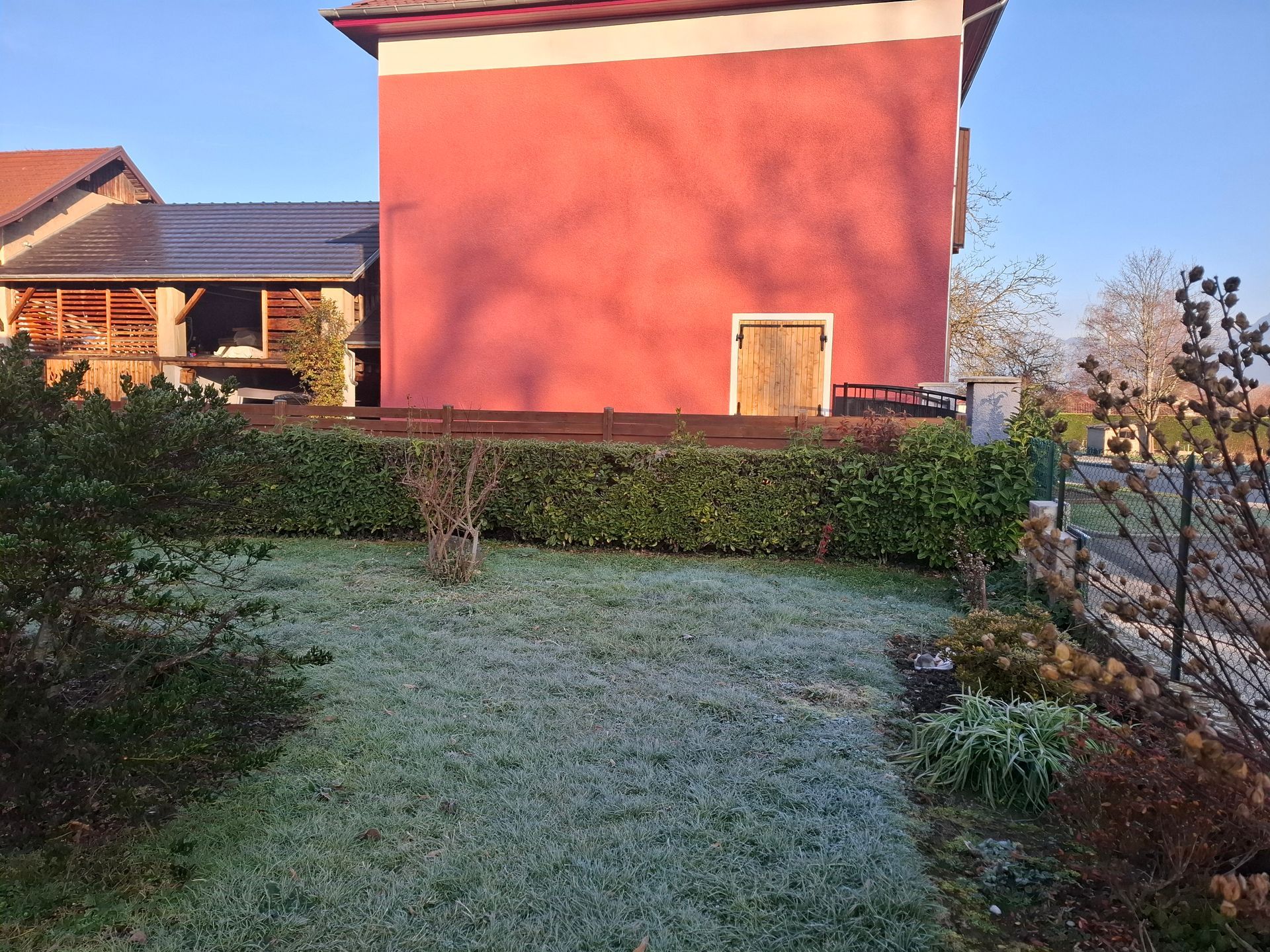 Une pelouse recouverte de givre devant un bâtiment rouge, une haie verte et un ciel bleu.
