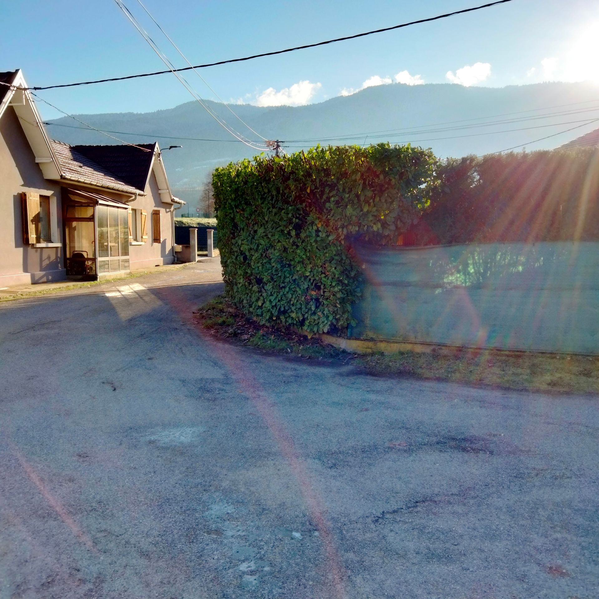 Une rue bordée de maisons, une haie et des montagnes sous un soleil éclatant.