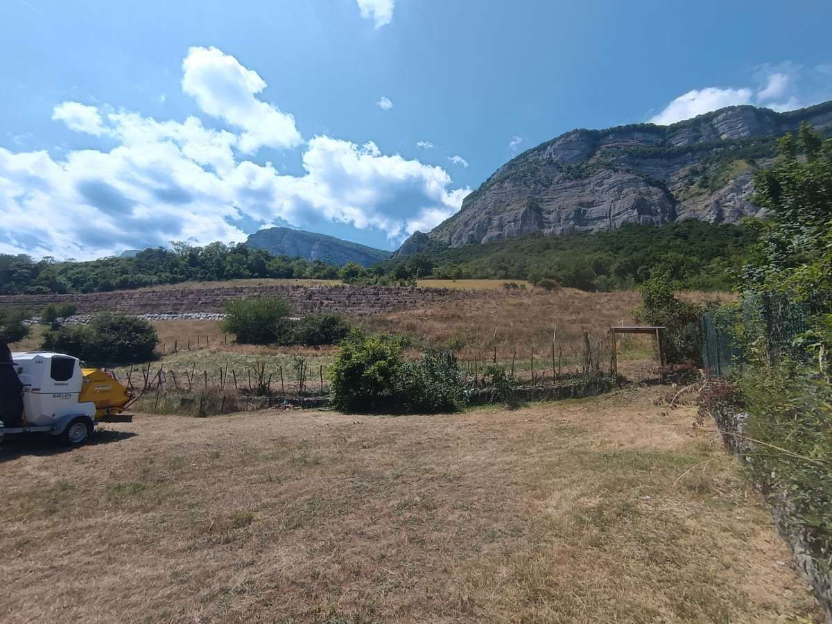 Champ herbeux avec des montagnes en arrière-plan sous un ciel bleu. On aperçoit quelques buissons, une clôture et un véhicule.