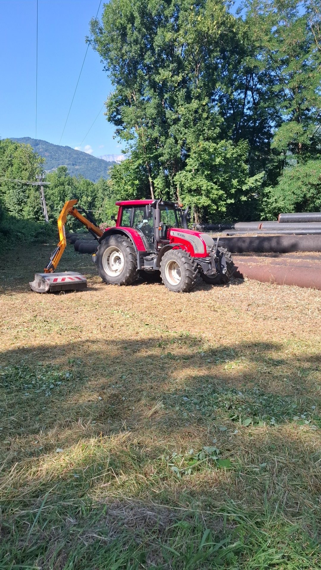 Tracteur rouge équipé d'un bras de coupe, coupant la végétation dans un champ avec des montagnes en arrière-plan.