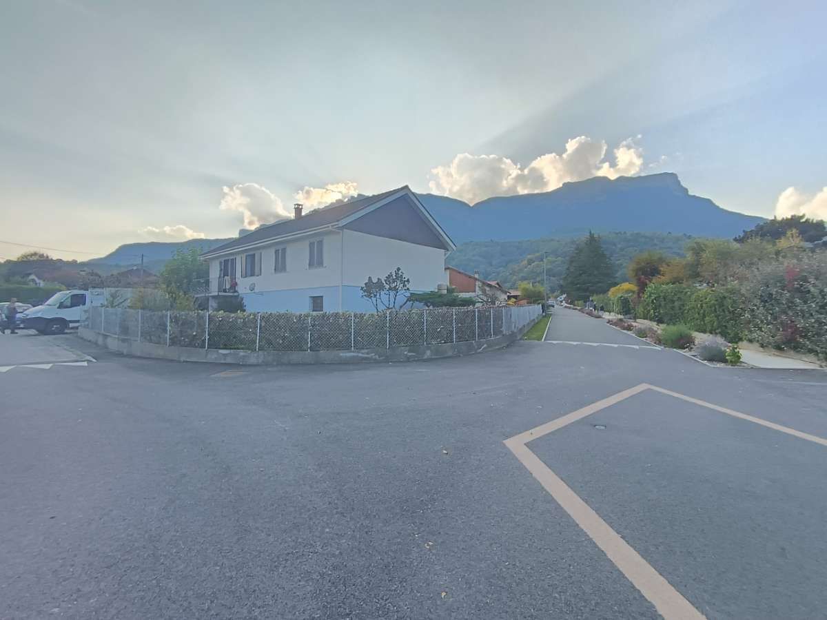 Maison en bord de route avec les montagnes en arrière-plan, ciel partiellement nuageux.