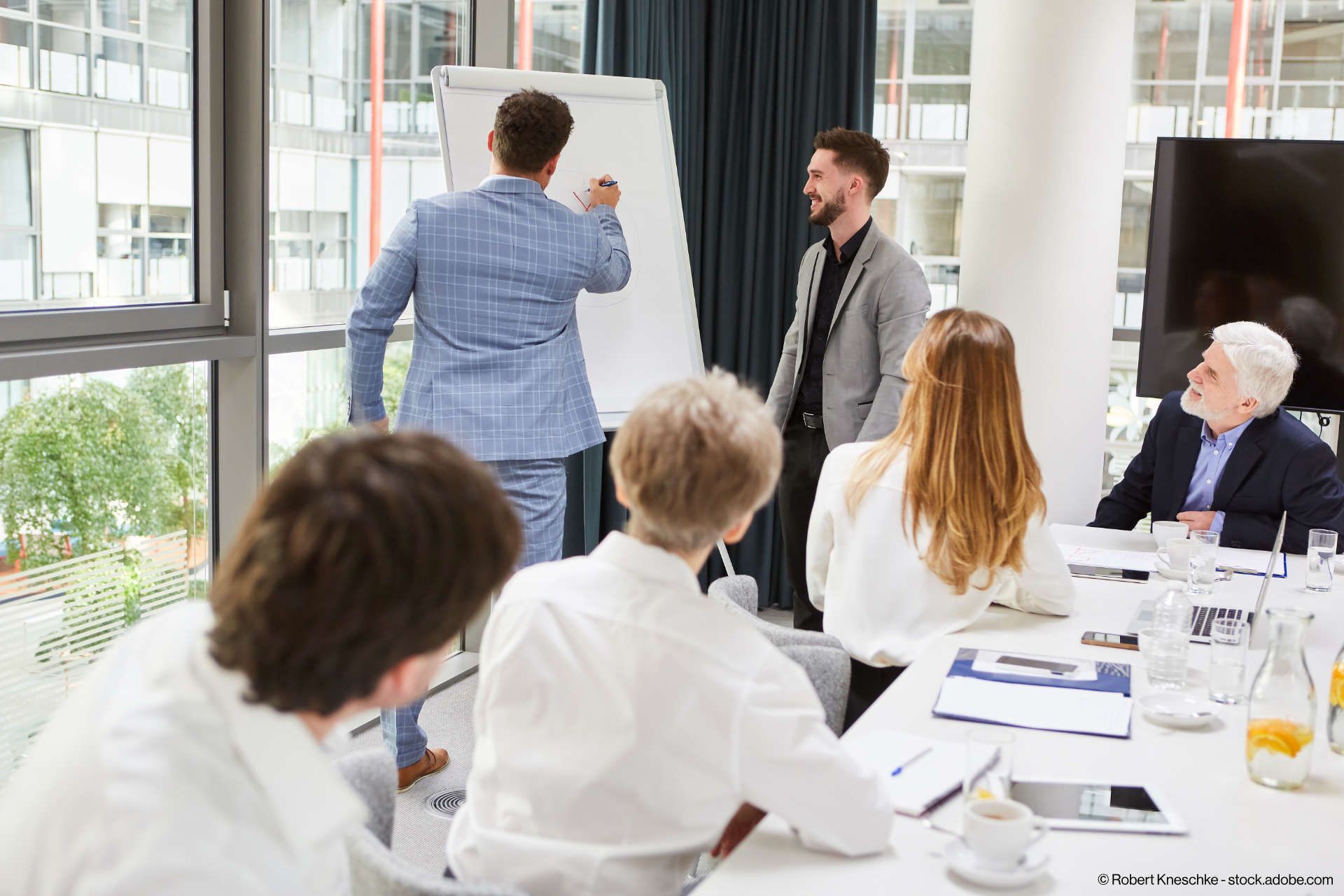 Ein Mann schreibt auf ein Flipchart, während eine Gruppe von Menschen um einen Tisch sitzt.