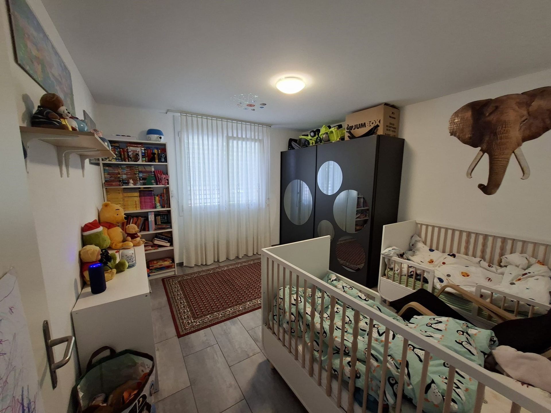 Camera da letto per bambini con culla, armadio, giocattoli, finestra e decorazione a forma di testa di cervo appesa al muro.