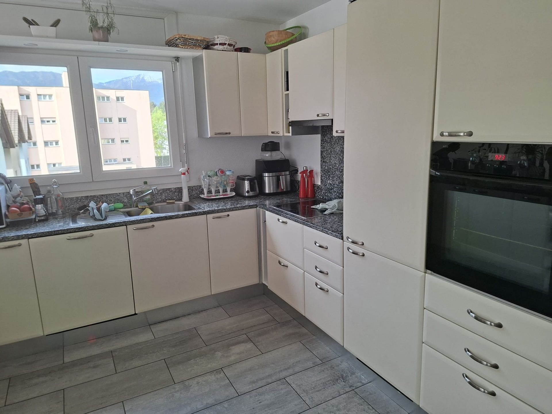 Cucina luminosa con mobili bianchi, piastrelle grigie sul pavimento e una finestra sopra il lavello.