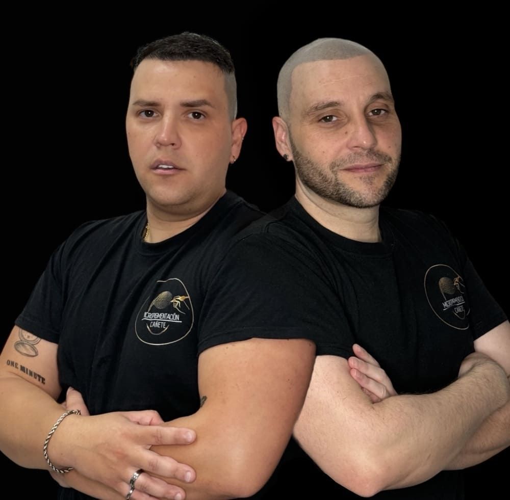 Dos hombres con camisas negras posan con los brazos cruzados, sobre un fondo negro.