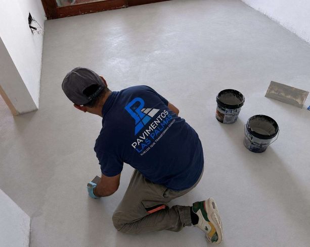 Un trabajador arrodillado sobre un suelo gris aplica una capa de pintura cerca de dos cubos de pintura y una llana.