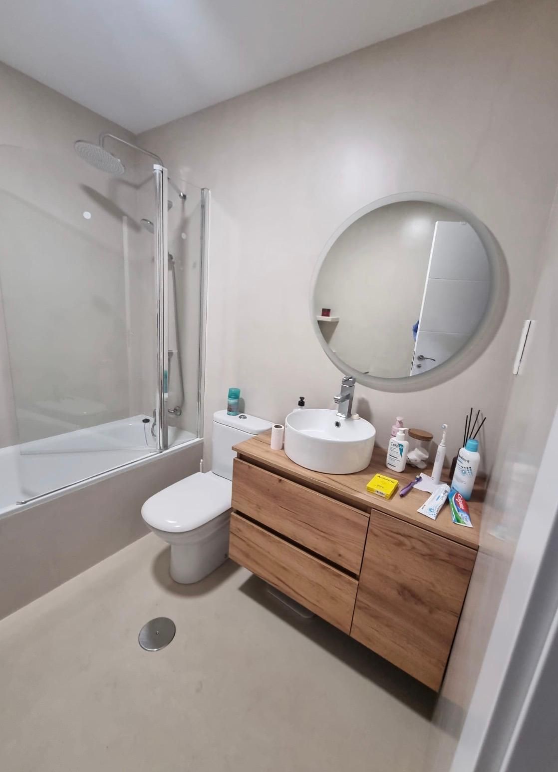 Baño pequeño con bañera, inodoro, espejo redondo, lavabo de madera y cortina de ducha.
