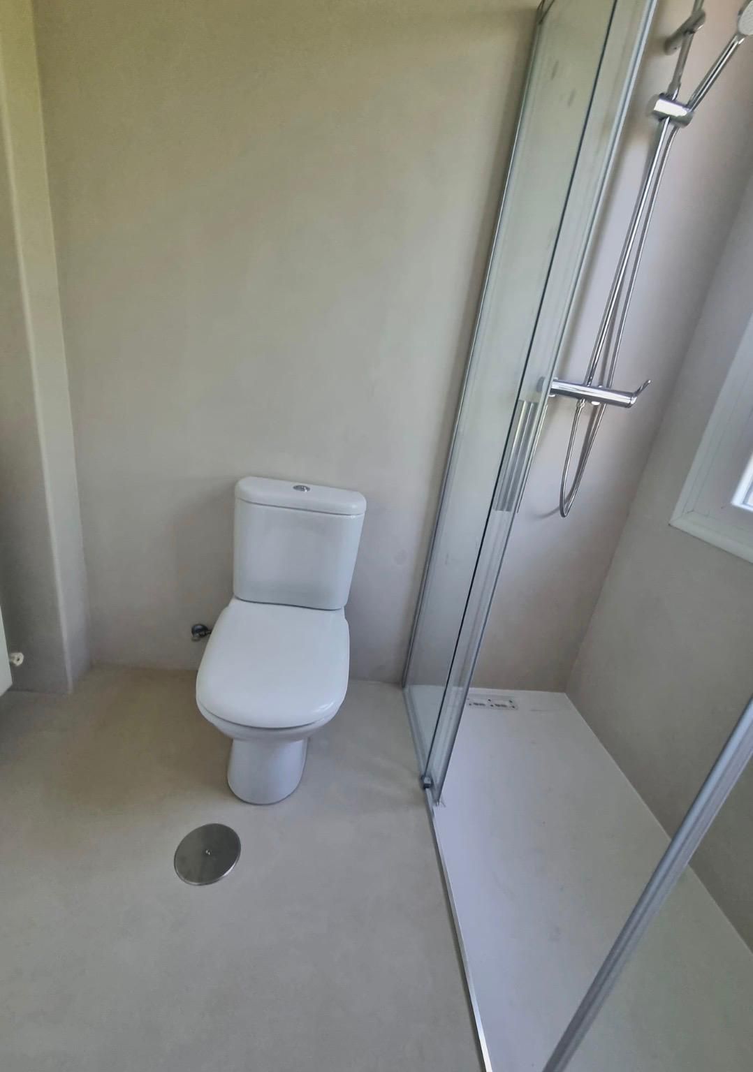Baño pequeño con inodoro blanco, desagüe en el suelo y cabina de ducha de cristal.