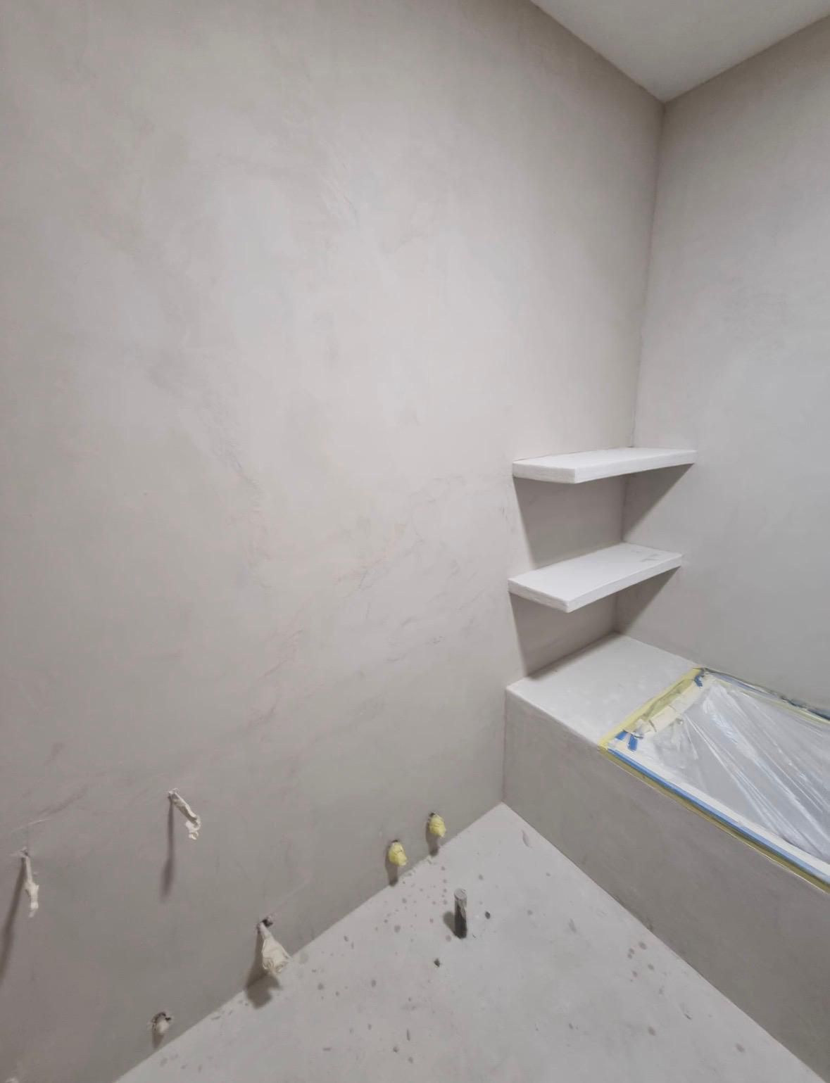 Habitación vacía sin terminar, con paredes blancas, estantes en las esquinas y una cama o colchón parcialmente cubierto.