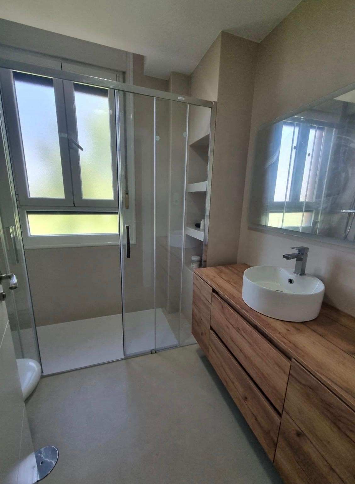 Baño moderno con ducha de cristal, tocador de madera y lavabo redondo junto a la ventana.