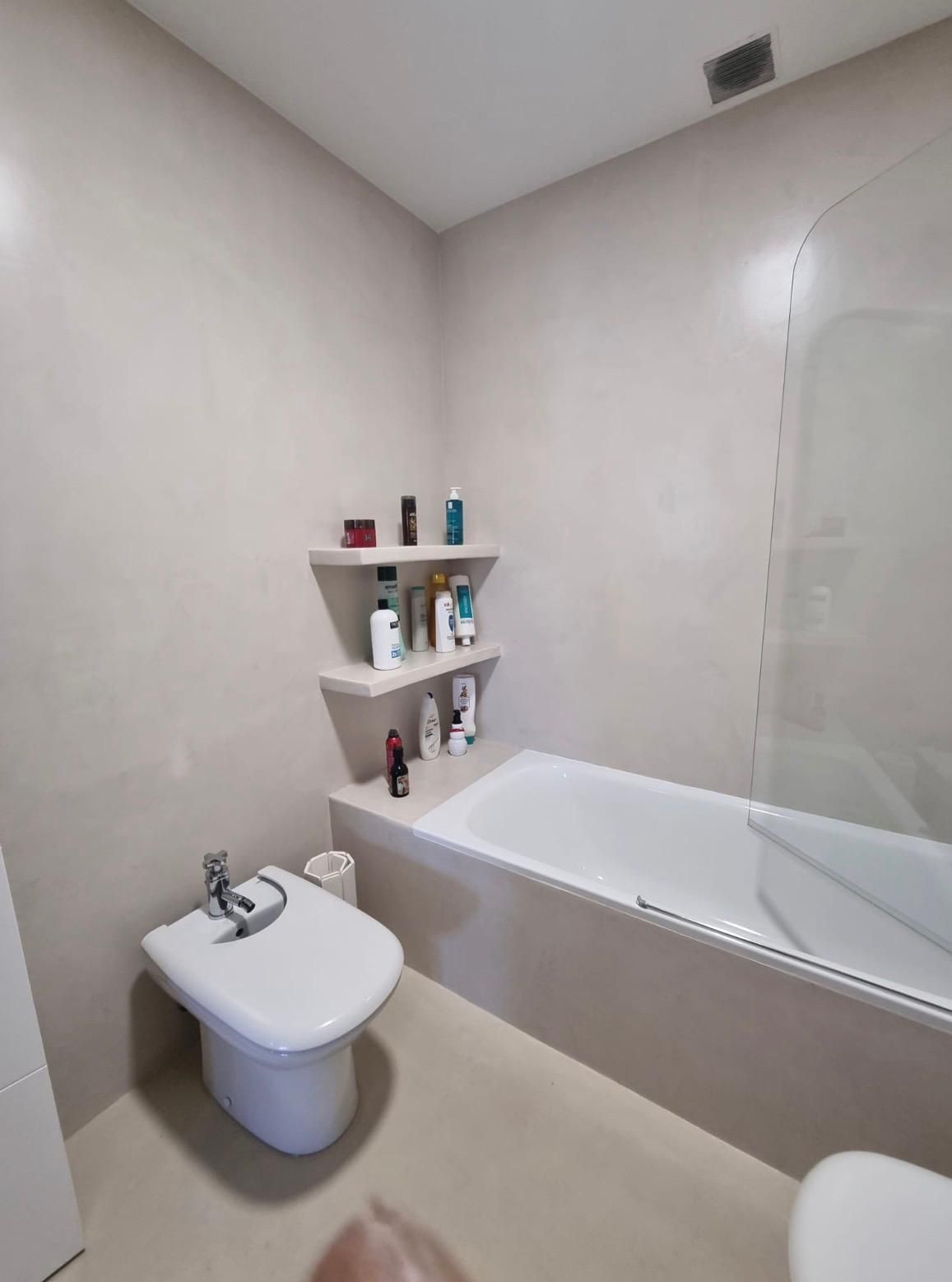 Pequeño baño de color beige con bañera, lavabo mural y estantes de esquina con artículos de aseo.