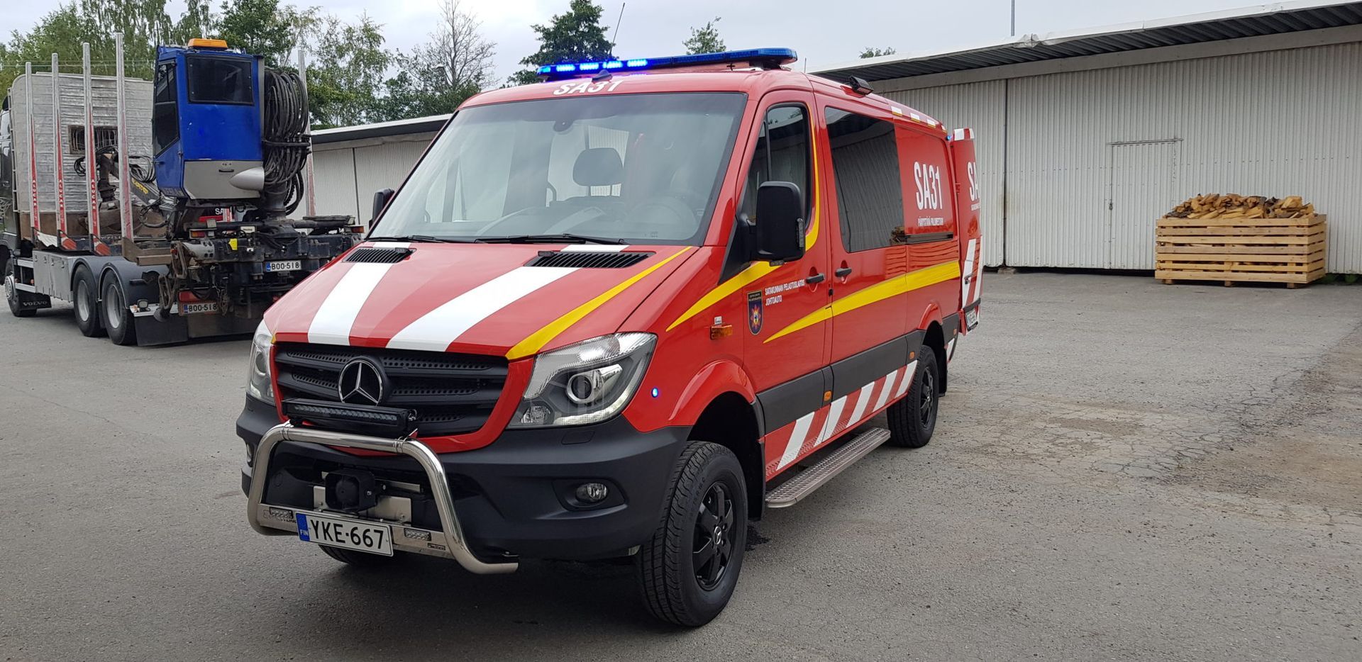 Punainen pelastusauto pysäköitynä soralle varaston viereen, taustalla kuorma-auto.