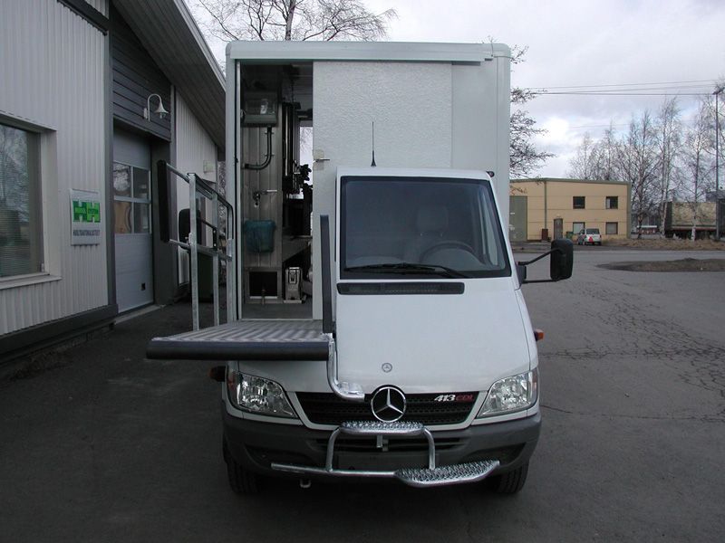Valkoinen Mercedes-jakeluauto, jossa on avoin sivustanostin, edestäpäin varaston ulkopuolella.