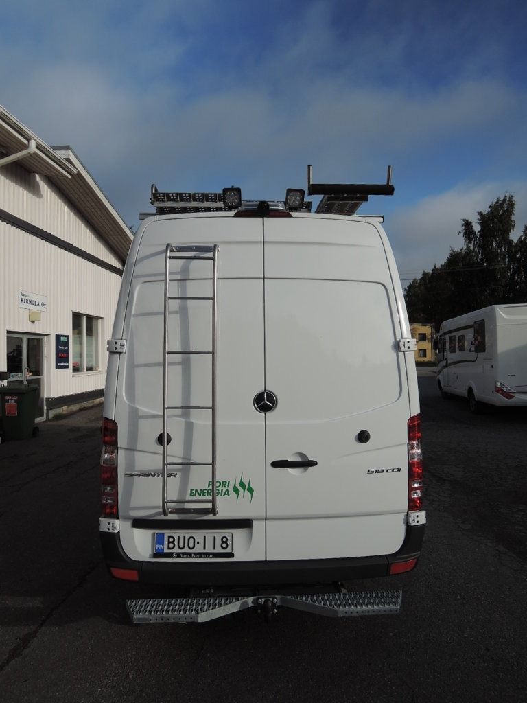Ulkona rakennusten vieressä pysäköidyn valkoisen pakettiauton takaa näkymä, jossa on kattotelineet ja tikkaat takaosassa.