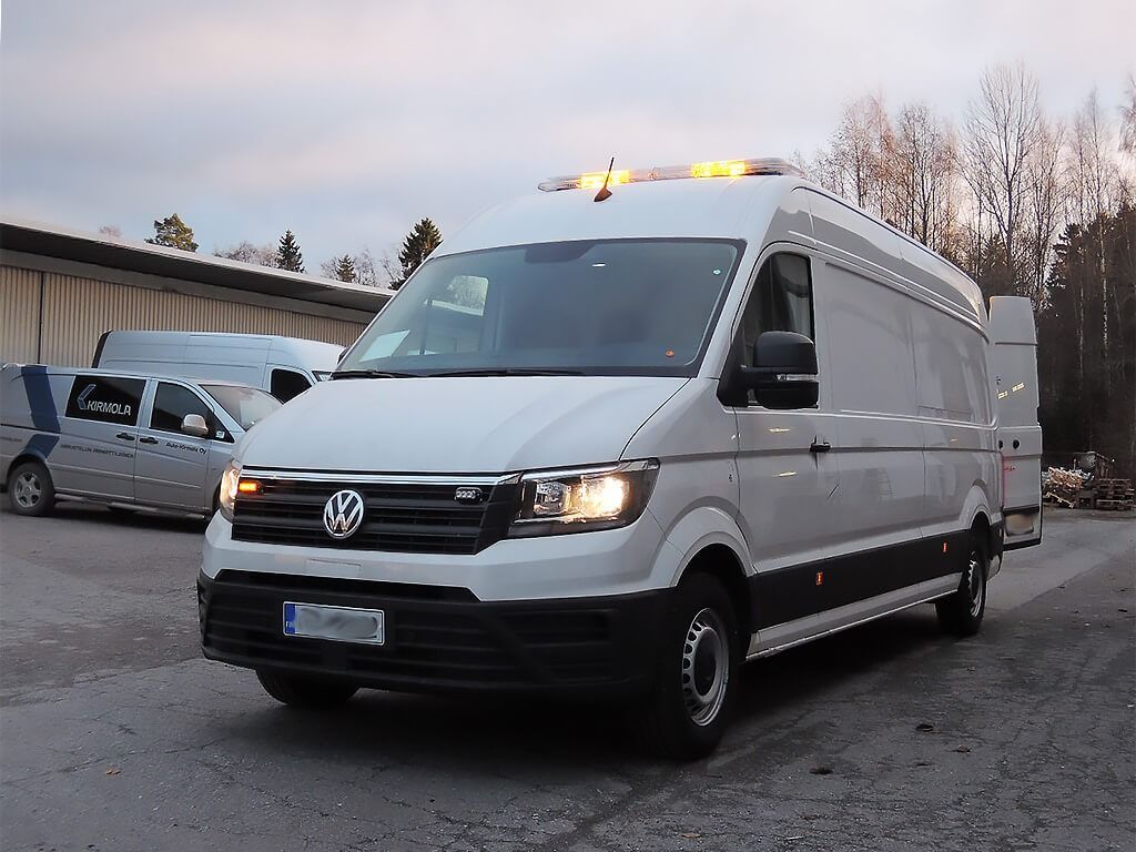 Valkoinen Volkswagen-pakettiauto keltaisilla kattovaloilla pysäköitynä lumiselle tontille teollisuusrakennusten lähelle