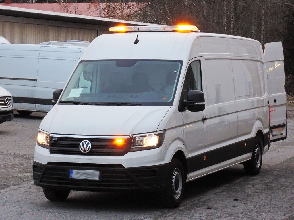 Valkoinen Volkswagen-pakettiauto, jossa on meripihkanväriset kattovalot, pysäköitynä ulkona märälle tontille