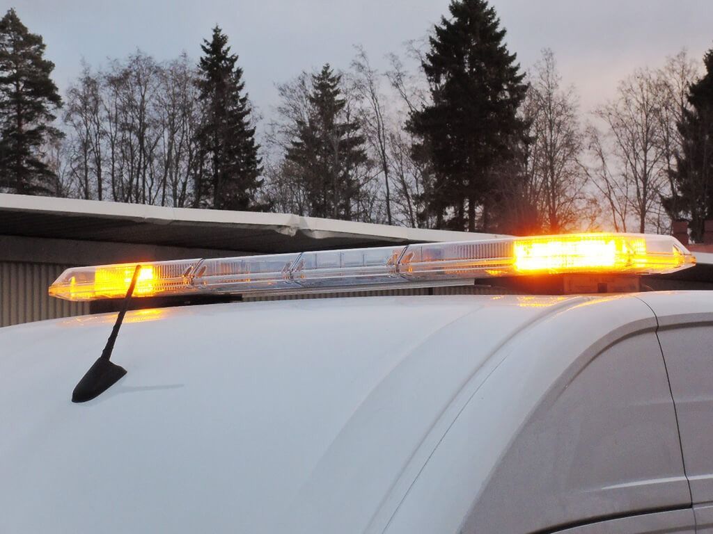 Keltainen hätävalopalkki valkoisessa ajoneuvossa hämärässä, taustalla puita.