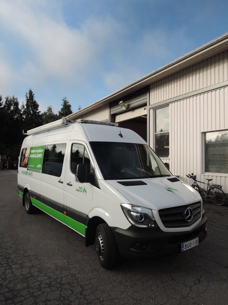 Valkoinen vihreäraitainen Mercedes-pakettiauto pysäköitynä teollisuusrakennuksen viereen soratietälle.