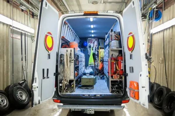 Avoin pakettiauto pysäköitynä korjaamoon, takaovet auki, työkalut ja laitteet näkyvät sisällä.