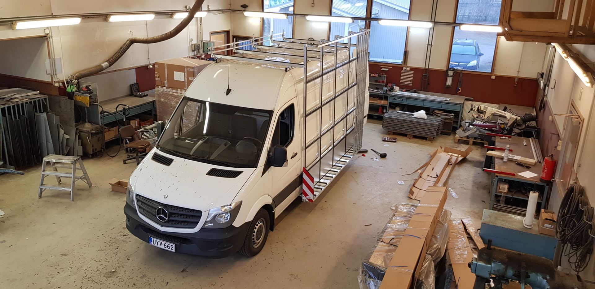 Valkoinen pakettiauto korjaamon autotallissa, työkalujen, tikkaiden ja työtasojen ympäröimänä.