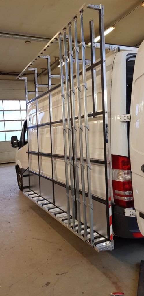 Valkoinen pakettiauto, jossa on korkea metallinen tavarateline, pysäköitynä autotalliin tai varastoon.