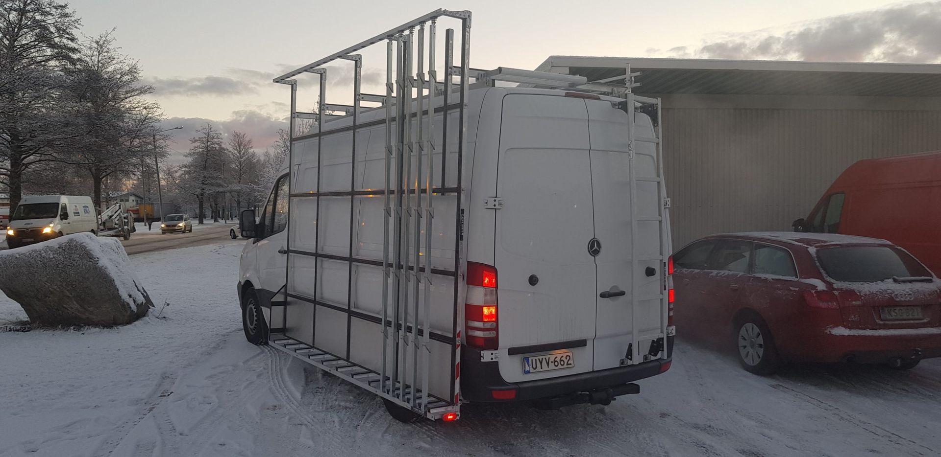 Valkoinen pakettiauto kattotelineellä ja jarruvaloilla pysäköitynä lumiselle kadulle punaisen auton viereen hämärässä