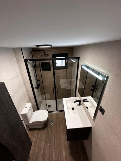 Baño moderno y compacto con ducha de cristal, espejo de pared, lavabo con mueble y retrete blanco.